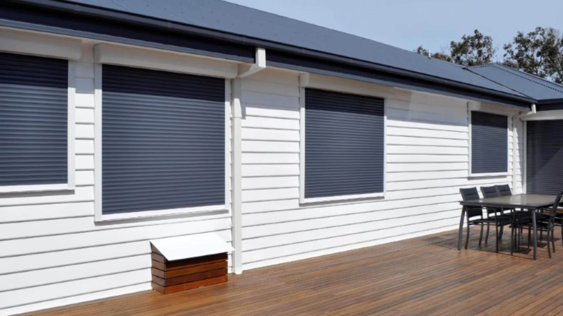 Several dark gray horizontal-slatted shutters cover windows along a white-sided house. The view runs diagonally along the facade beneath a dark roof, with the nearest window taking up much of the left and center. A wooden deck extends along the wall and holds a dark outdoor table with matching chairs. No people or readable text are visible.