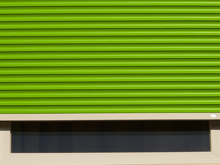 A bright green horizontally slatted roller shutter fills most of the image above a dark, long rectangular window. The shutter is lowered close to the window opening, with a white frame visible along the bottom and sides. A small white oval mark appears near the lower-right corner of the shutter. No people or readable text are visible.