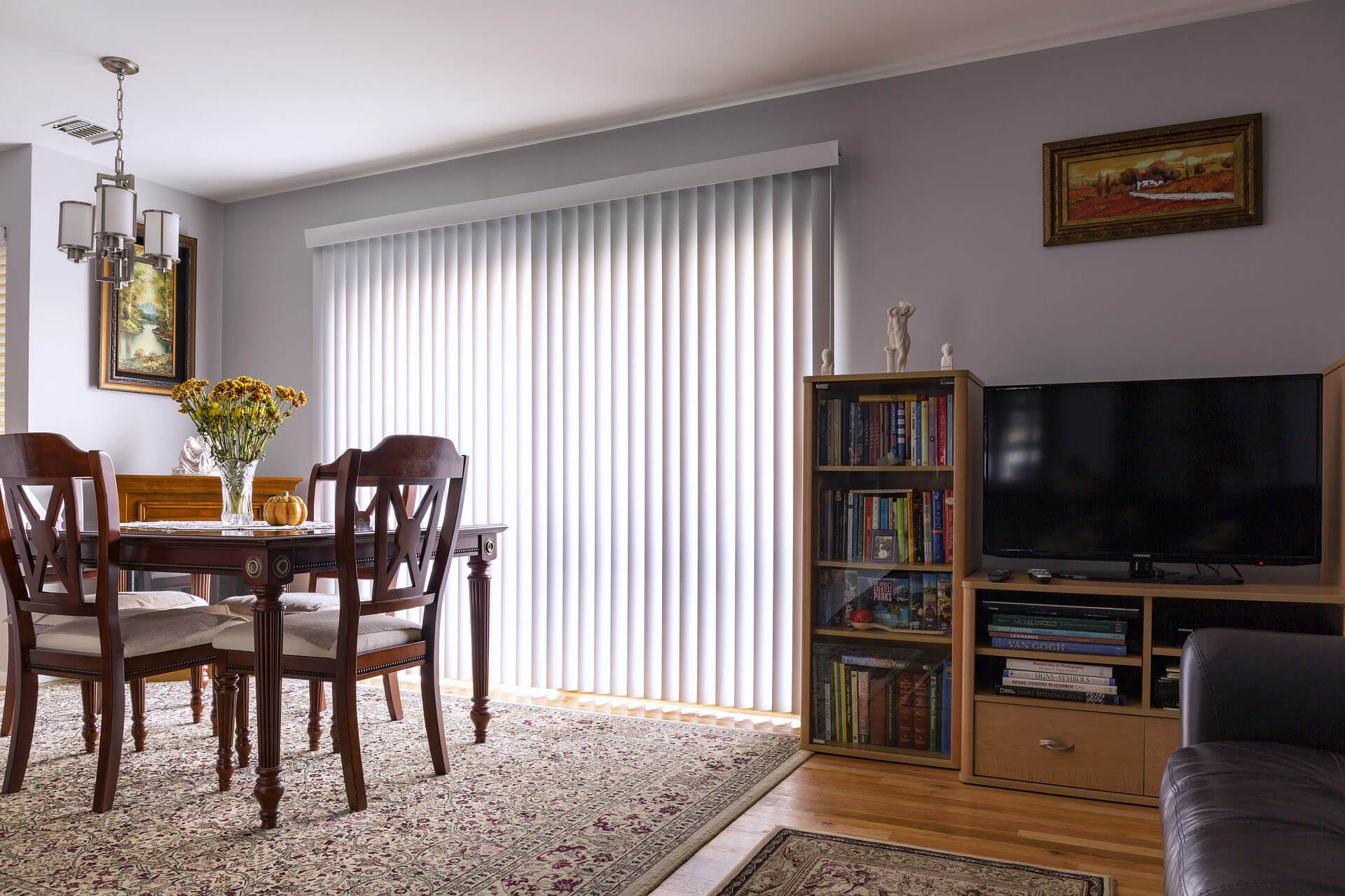 A wide interior view shows a dining area and a television cabinet in the same room. Tall white vertical fabric slats cover a large central window, with daylight visible between them. In the foreground are a dark wooden dining table and chairs, flowers, and patterned rugs; shelving, books, a television, framed art, and small statues are also visible. No people or readable text appear.