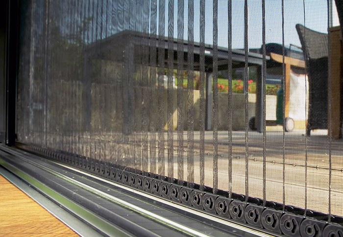 A dark fine-mesh panel spans most of the frame, with an outdoor paved area and building surfaces visible behind it. Along the bottom, the panel meets a metal track containing a row of round dark fittings. A warm-toned wooden surface enters at the lower left, and part of a chair is visible at the upper right. No people or readable text are visible.