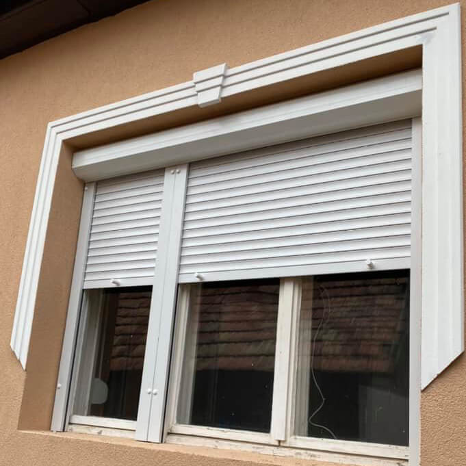 A white-framed window in a tan stucco wall is partly covered by a white horizontal-slatted shutter lowered over the top portion. The uncovered lower glass is divided into vertical sections and reflects a dark tiled roof. A projecting white surround frames the opening, with a small raised decorative detail centered above it. No people or readable text are visible.