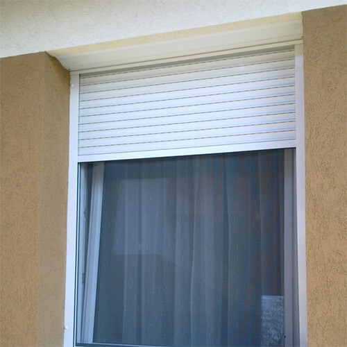 A white slatted shutter is partially lowered over the upper part of a window set into a beige exterior wall. The uncovered lower glass appears dark and has a sheer curtain behind it. White side tracks frame the shutter and window, with the image tightly cropped around the opening. No people or readable text are visible.