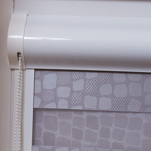 A white rounded roller cassette spans the top of a pale gray fabric shade. The shade has a repeating geometric pattern of rounded squares and textured inserts, with a horizontal seam or band across the lower-middle area. A white beaded pull chain hangs at the left beside the cassette end cap. The close crop shows the hardware and fabric against a pale wall.
