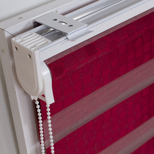 A close detail shows red patterned fabric arranged in wide horizontal bands across a window shade. A white roller mechanism and rectangular metal mounting bracket run along the top left, with parallel white rails extending across the top. Two strands of white beads hang beside the side bracket. The crop concentrates on the blind assembly against a pale background.