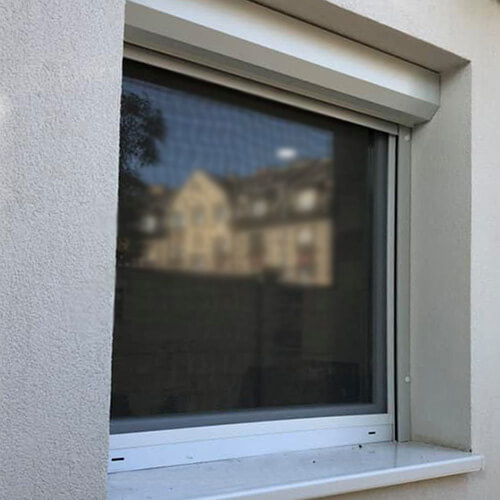 A close crop shows a rectangular window in a white textured exterior wall. A broad white housing and vertical side edges surround the top and sides of the opening. The dark window glass reflects a neighboring house and trees, with no visible slats across the glass. A white sill projects below; no people or readable text are visible.