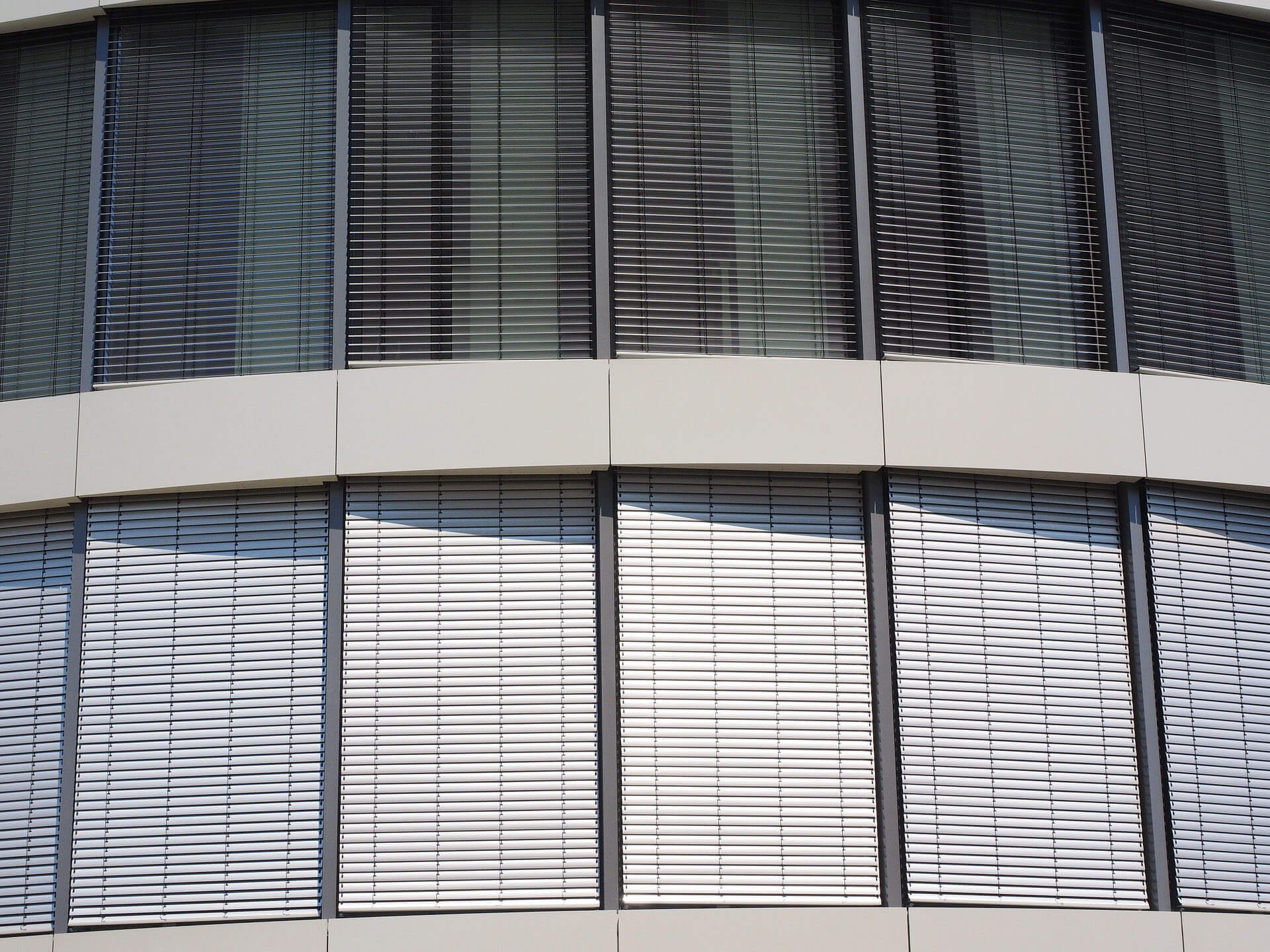A modern building facade fills the frame with rows of large windows fitted with closely spaced horizontal slats. The upper windows are shaded and dark, while lower windows admit bright light between the slats. Pale horizontal facade panels separate the upper and lower window bands. No people or readable text are visible.