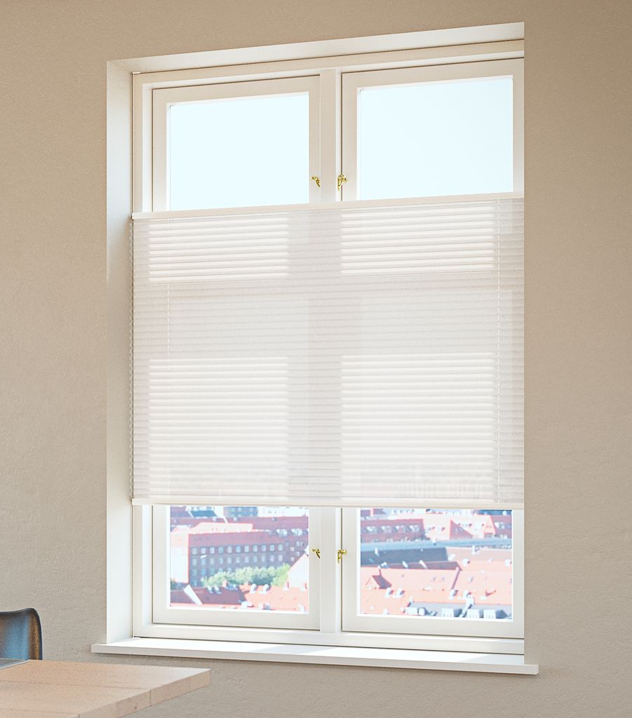 A white window covering made of narrow horizontal lines spans the middle of a tall double window and is positioned partway down. Fine repeated bands form an even texture across the material, with bright light passing through it. The uncovered upper and lower panes reveal a distant view of rooftops and buildings. Part of a pale tabletop and a chair edge appear in the lower left.