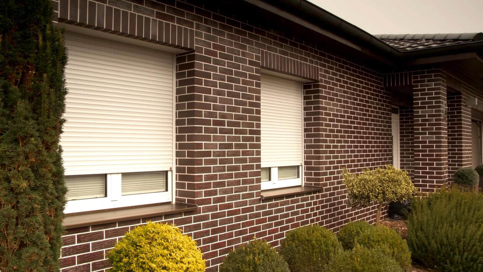 The image shows the exterior of a brick-patterned house with multiple white horizontal-slatted shutters lowered over windows. The nearest large shutter covers most of a wide window, while smaller sections of glazing remain visible near the lower edges. Shrubs and trimmed plants line the wall in the foreground. Roof eaves and a tiled roof are visible above; no people or readable text appear.