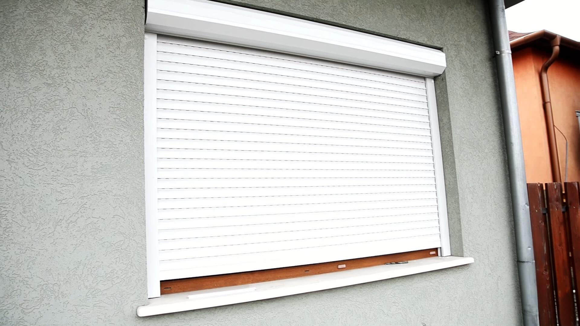 A close exterior view shows a white horizontally slatted shutter lowered over a window in a gray textured wall. Most of the opening is covered, leaving a narrow strip of brown sill visible at the bottom. A white projecting window ledge runs across the foreground. At the right edge are a downspout and part of an orange wall with a wooden fence; no people or readable text appear.