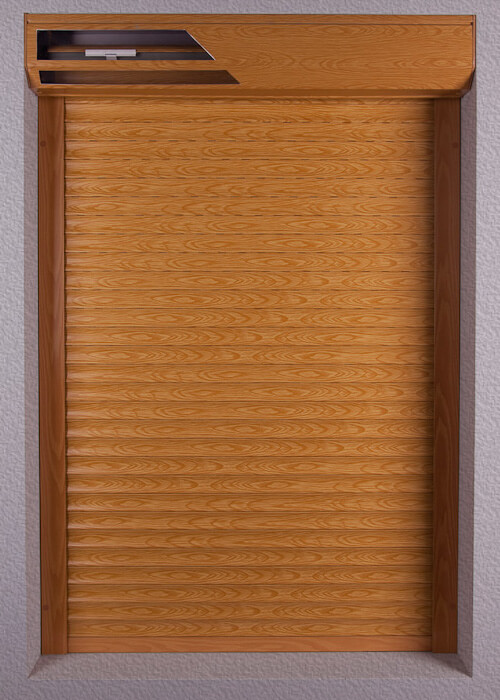 A rectangular shutter is shown closed against a light gray textured wall. Its horizontal slats and the surrounding top housing and side rails have a warm brown wood-grain appearance. A narrow dark opening and a small pale component are visible in the upper housing.