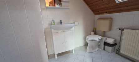 Tetőtéri fürdőszoba mosdóval, WC-vel, radiátorral és ferde fa mennyezettel.