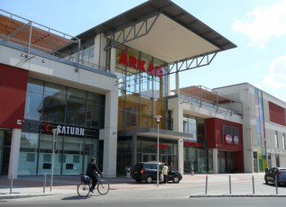 Glass-fronted ÁRKÁD shopping center entrance with a cyclist and parked cars.