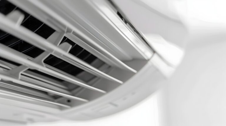 The image is a close-up view into the lower outlet of a white air-conditioning unit. Pale horizontal louvers and dark internal fins fill the left and center of the frame, while the right side falls into bright soft focus. The composition emphasizes the machine's internal airflow opening rather than a full room or product view.