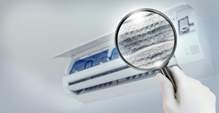 A wall-mounted air-conditioning unit is shown out of focus in the background with its front panel open. In the foreground, a gloved hand holds a magnifying glass over a visibly dusty filter surface. The cool blue-gray image uses the magnifier and filter texture as the main focal point.