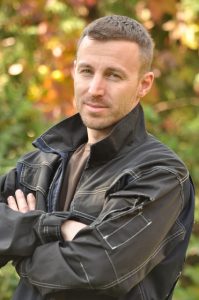 Man in a black leather jacket standing outdoors with folded arms.
