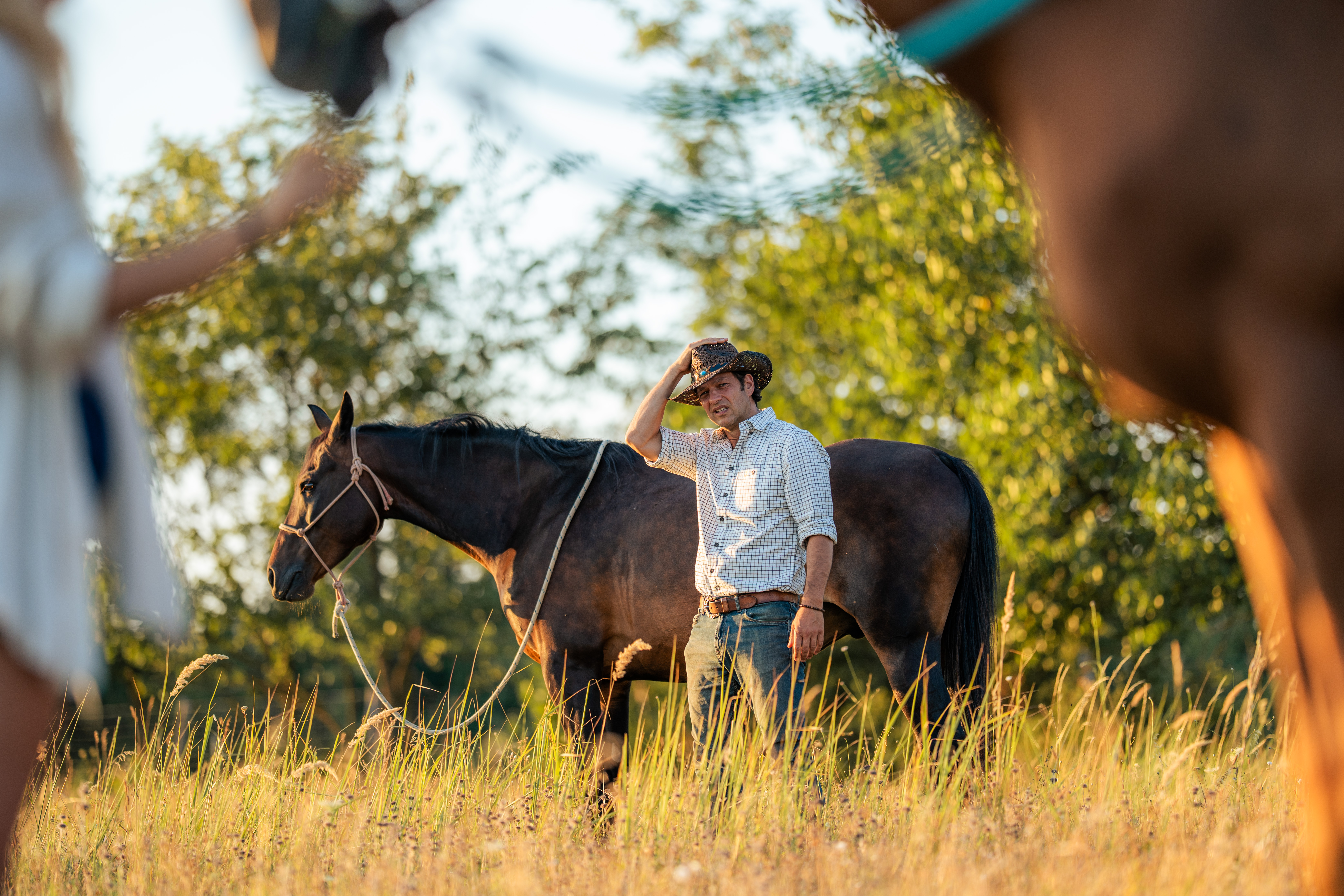 A person wearing a wide-brimmed woven hat and checked shirt stands beside a dark horse. One hand is raised to the hat brim, and the horse's head is lowered at the left. Blurred shapes at both sides frame the subjects, with green foliage bokeh and tall grass behind them. No text is visible.