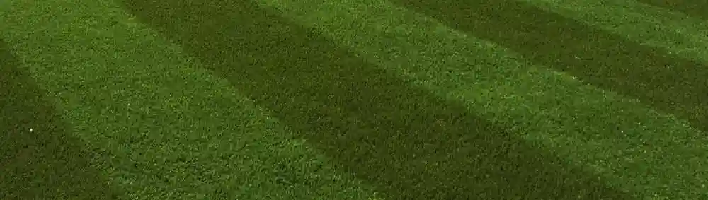 The frame is filled by a close view of dense short green grass. The surface has subtle darker and lighter bands running diagonally across the image. No buildings, people, or tools are visible.