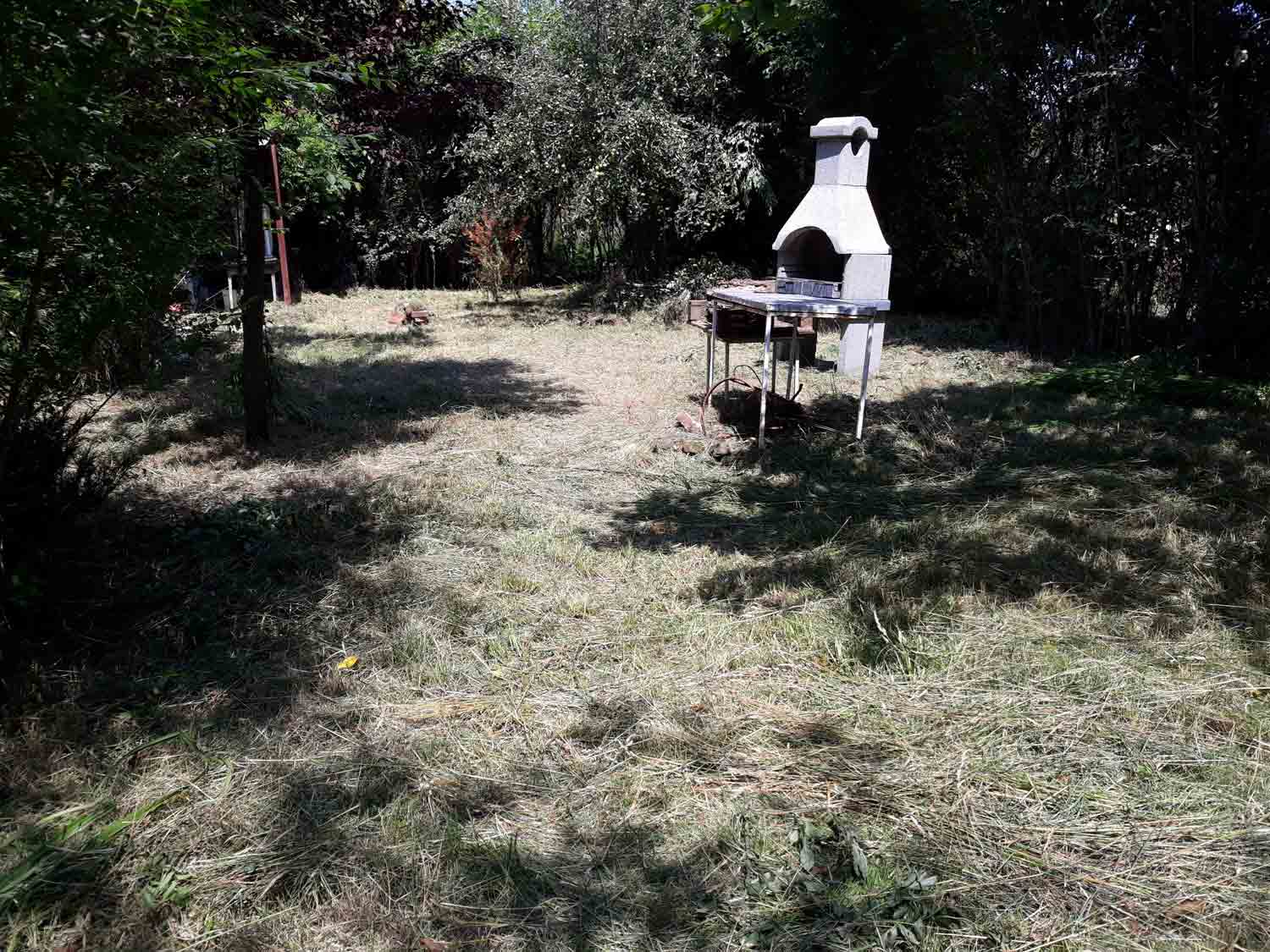 A cleared garden area is covered with short, dry grass and scattered cut vegetation. A white masonry structure with an arched opening stands on the right beside a metal-topped table. Dense trees and shrubs border the space.