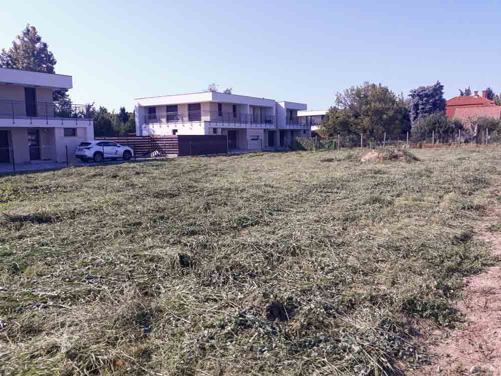 The foreground is a wide field of short, cut grass with a bare dirt strip at the lower right. White modern houses and a parked white car are visible behind the field. Trees, fencing, and a red-roofed building form the background under a pale blue sky.