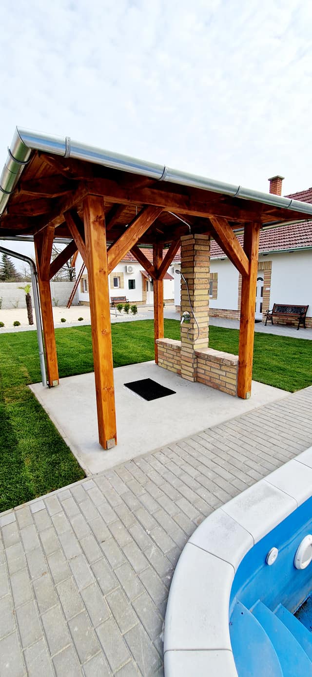 A fa pergola mellett a medence kék pereme és kövezett járda látható.