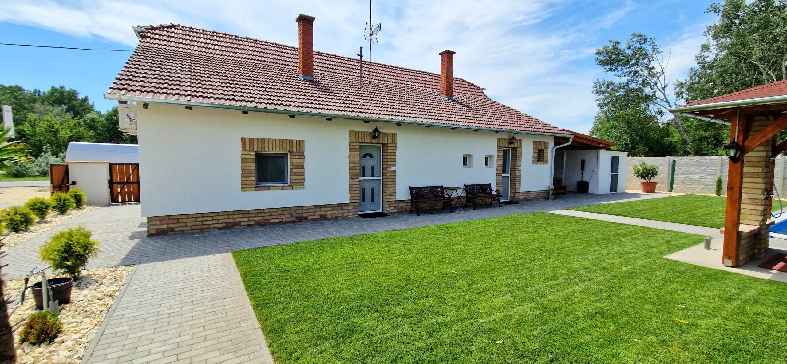 A fehér, alacsony épület előtt széles nyírt gyep és fedett fa pavilon látható.