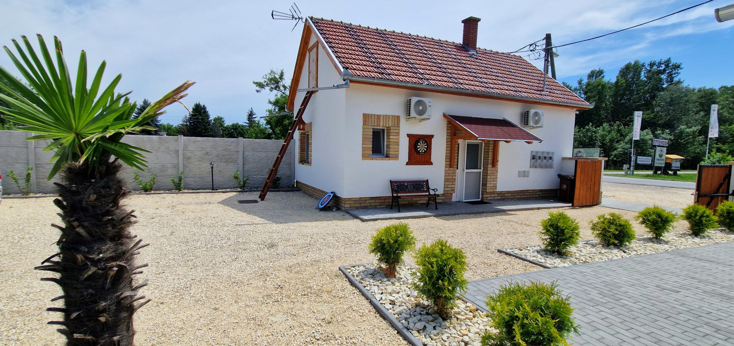 A kavicsos udvar közepén kis fehér, piros tetős épület és pálmafa áll.