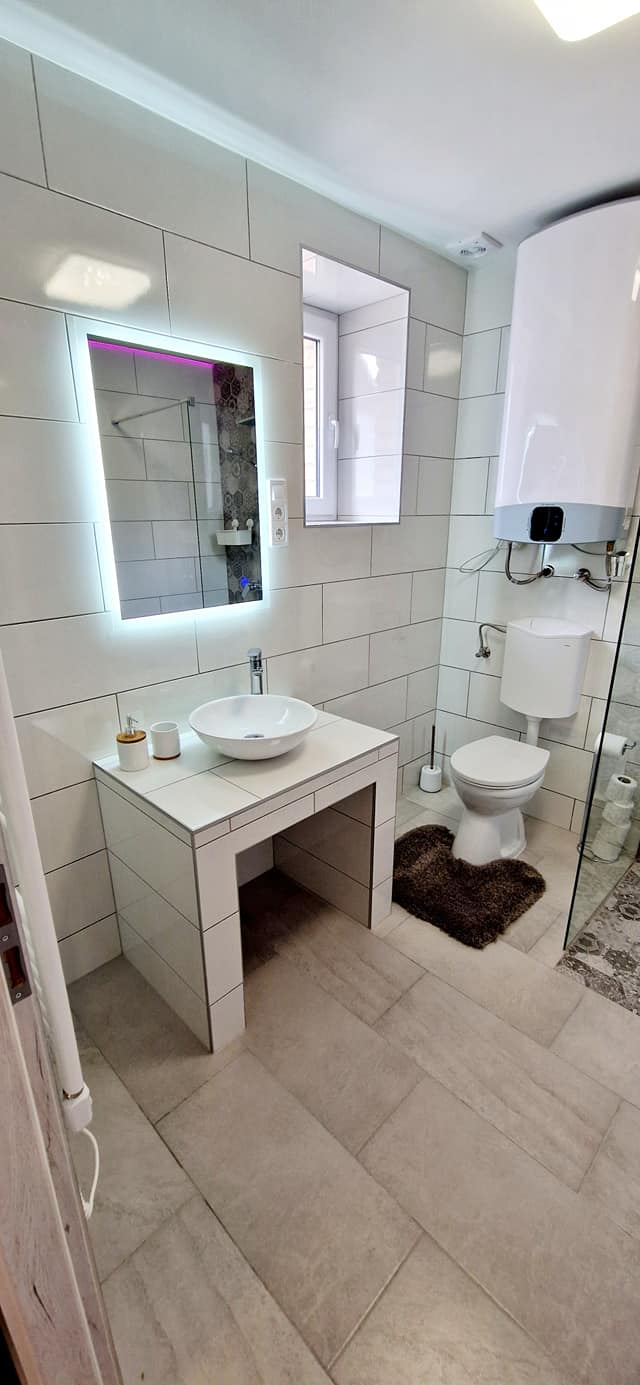A keskeny helyiségben mosdó, tükör és zuhanyzó látható.