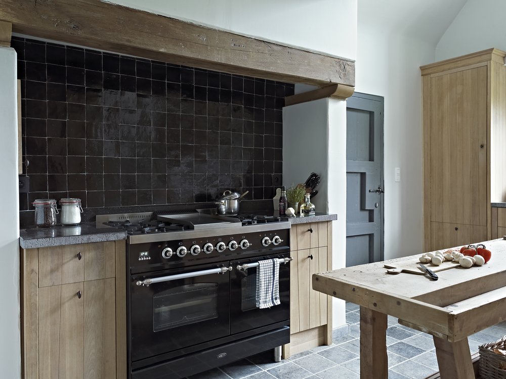 Dark tile backsplash, black range and tall pale wood cabinet by a window.