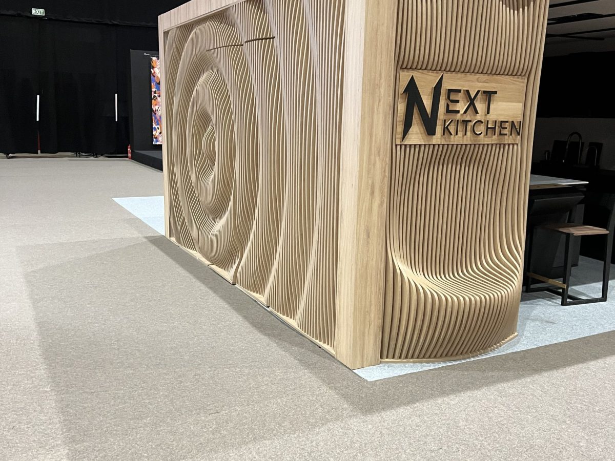 Curved pale wood display wall with next kitchen logo at the corner.